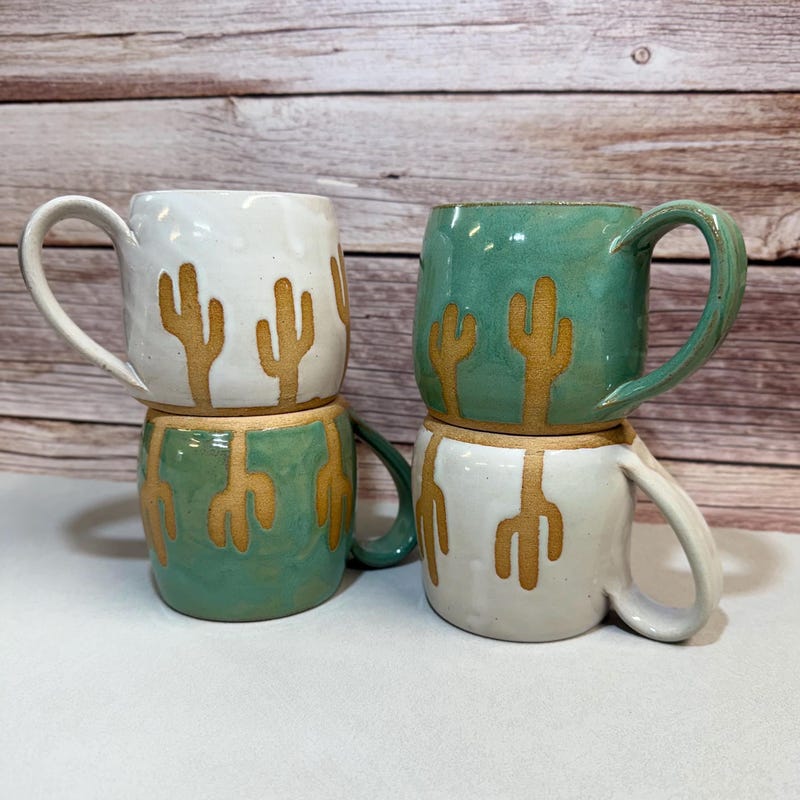 Four cactus mugs stacked in pairs, cream and sea green with saguaro shapes left in bare clay.
