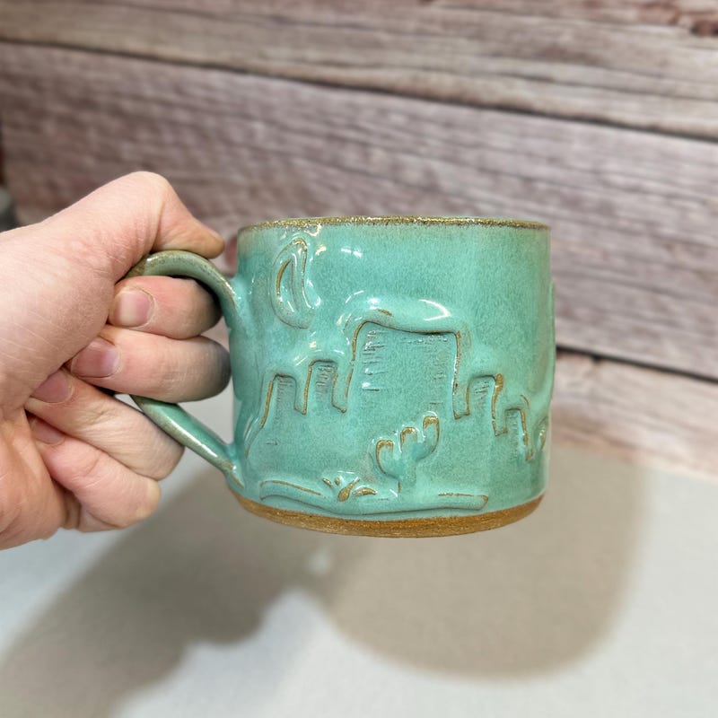 Sea-green mug carved with a desert cactus scene and a bare clay rim, held in one hand.