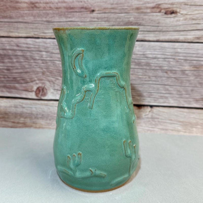 Sea-green vase carved with saguaro cactus and a crescent moon, against weathered barn wood.