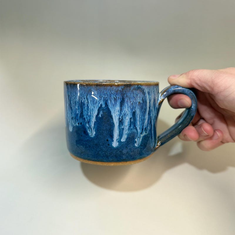Short deep blue cappuccino mug with white glaze dripping from the rim, held in one hand.
