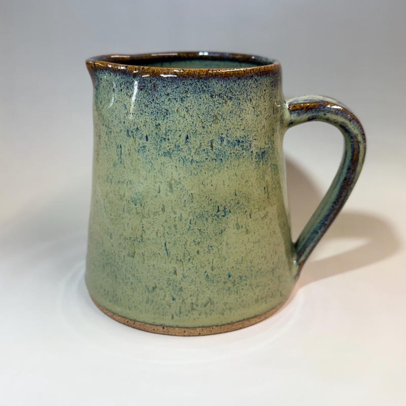 Sage green pitcher with a speckled glaze, a pulled handle and a bare clay foot.