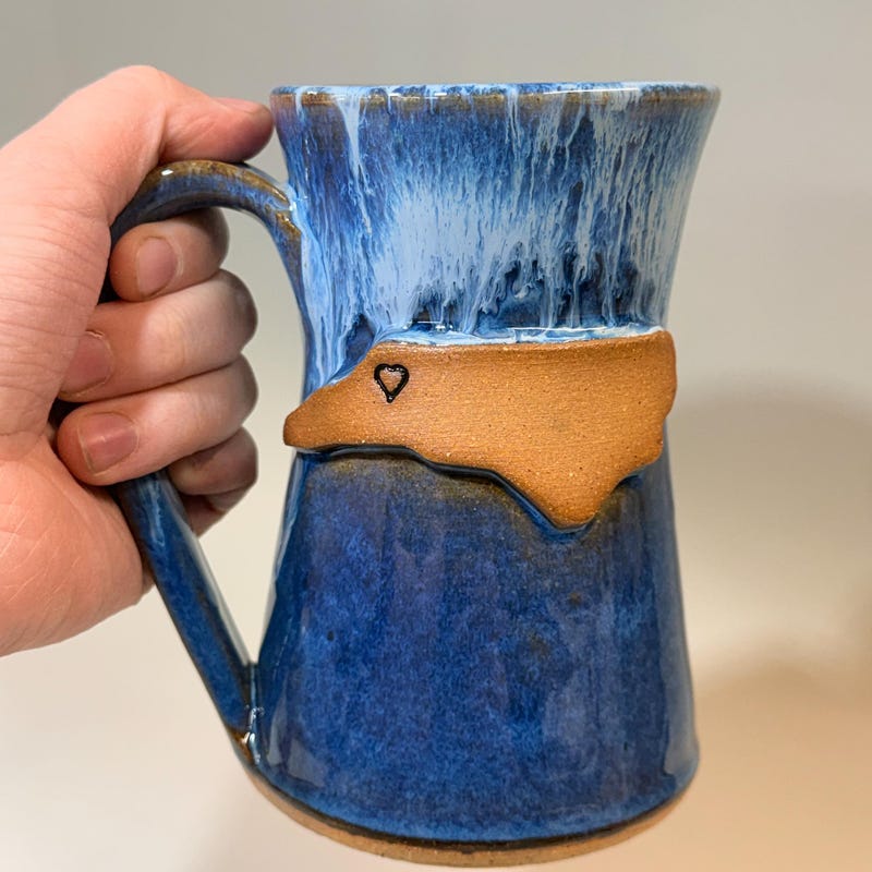 Blue mug held in one hand, the outline of North Carolina in bare clay with a small heart stamped into it.