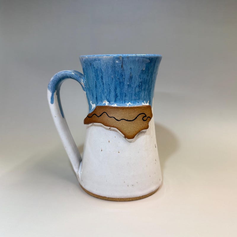 White and blue mug with the outline of North Carolina in bare clay, drawn with a mountain and a wave.