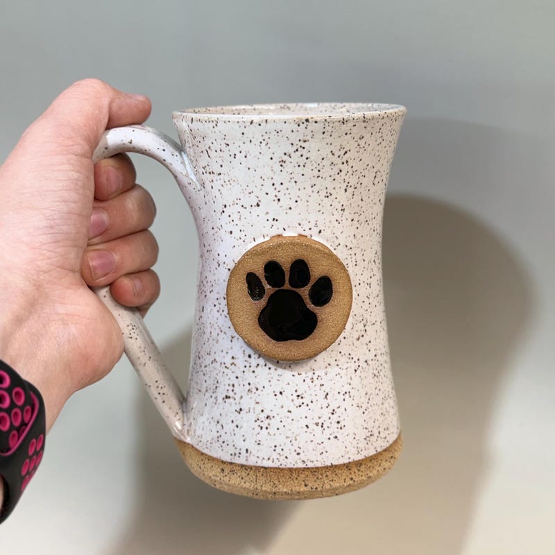 Speckled white mug held in one hand, a black paw print set into a circle of bare clay.
