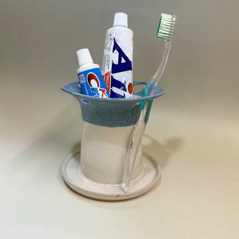 Cream and pale blue toothbrush holder on a wide saucer base, holding a toothbrush and two tubes of toothpaste.