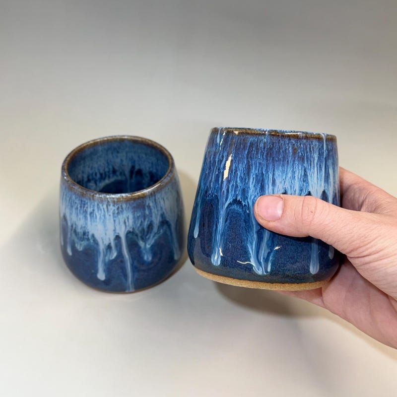 Two handleless blue tumblers with white glaze running down the sides, one of them held in a hand.