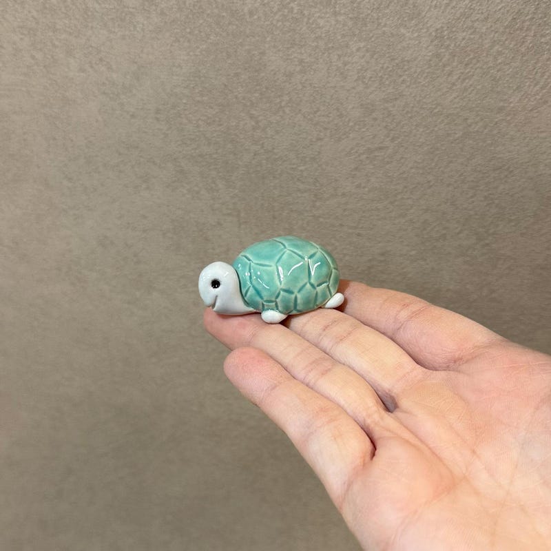 A miniature ceramic turtle with a turquoise shell, sitting in the palm of a hand.